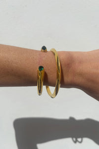 A close-up of a person's wrist wearing three gold bangle bracelets, including one with a small green gemstone and one with a small dark gemstone, against a plain white wall.