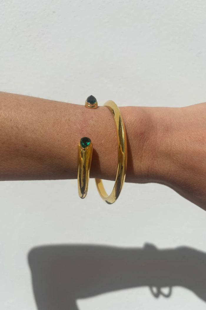 A close-up of a person's wrist wearing three gold bangle bracelets, including one with a small green gemstone and one with a small dark gemstone, against a plain white wall.