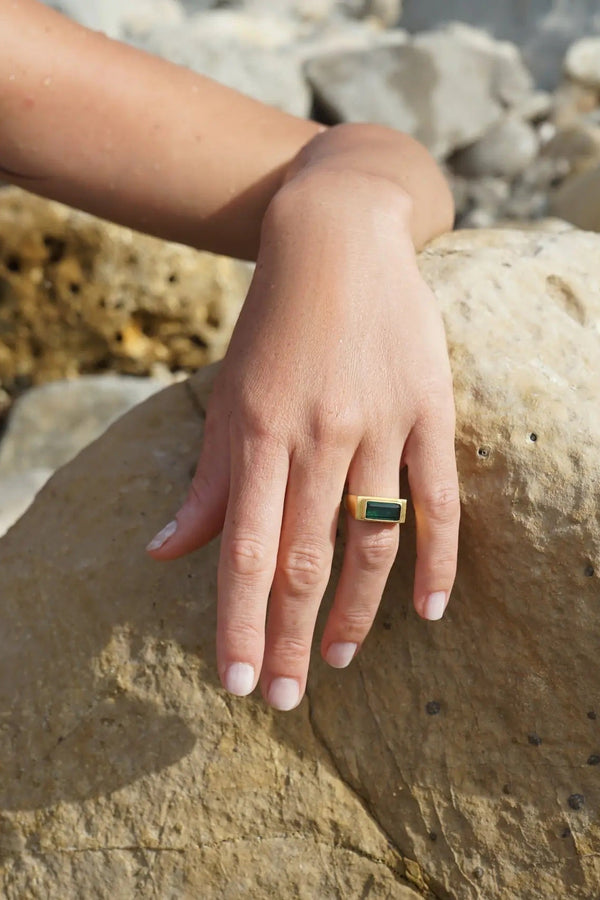 A gold ring with a rectangular green stone worn on a woman's ring finger, shown in a lifestyle setting on a rocky beach.