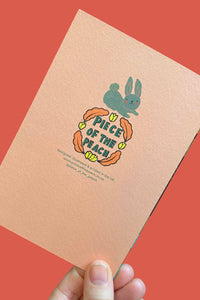 A hand holds a peach-colored notebook or card featuring a cute illustration of a blue bunny inside a peach outline, with the text 'Piece of the Peach' in yellow and green. The background is a solid coral-red color.