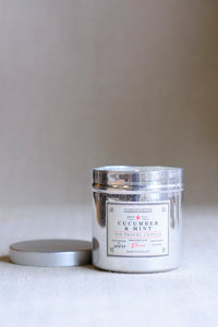 A silver metal tin candle labeled 'FARMLANDS Cucumber & Mint Tin Travel Candle' with its matching silver lid resting beside it on a neutral beige surface.