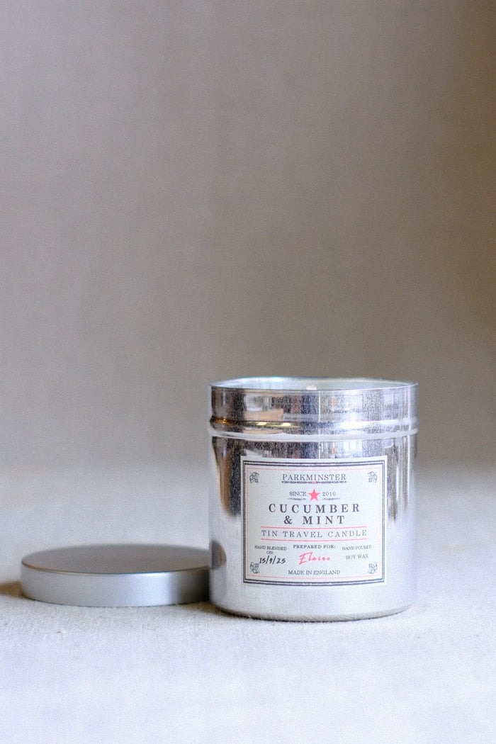 A silver metal tin candle labeled 'FARMLANDS Cucumber & Mint Tin Travel Candle' with its matching silver lid resting beside it on a neutral beige surface.
