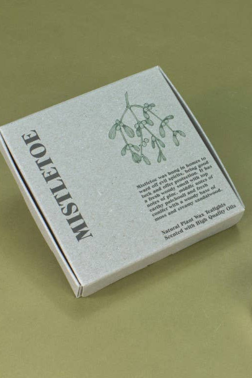 A square gray box labeled 'MISTLETOE' with a botanical illustration and product text, shown beside three small white tea light candles on a muted olive background.
