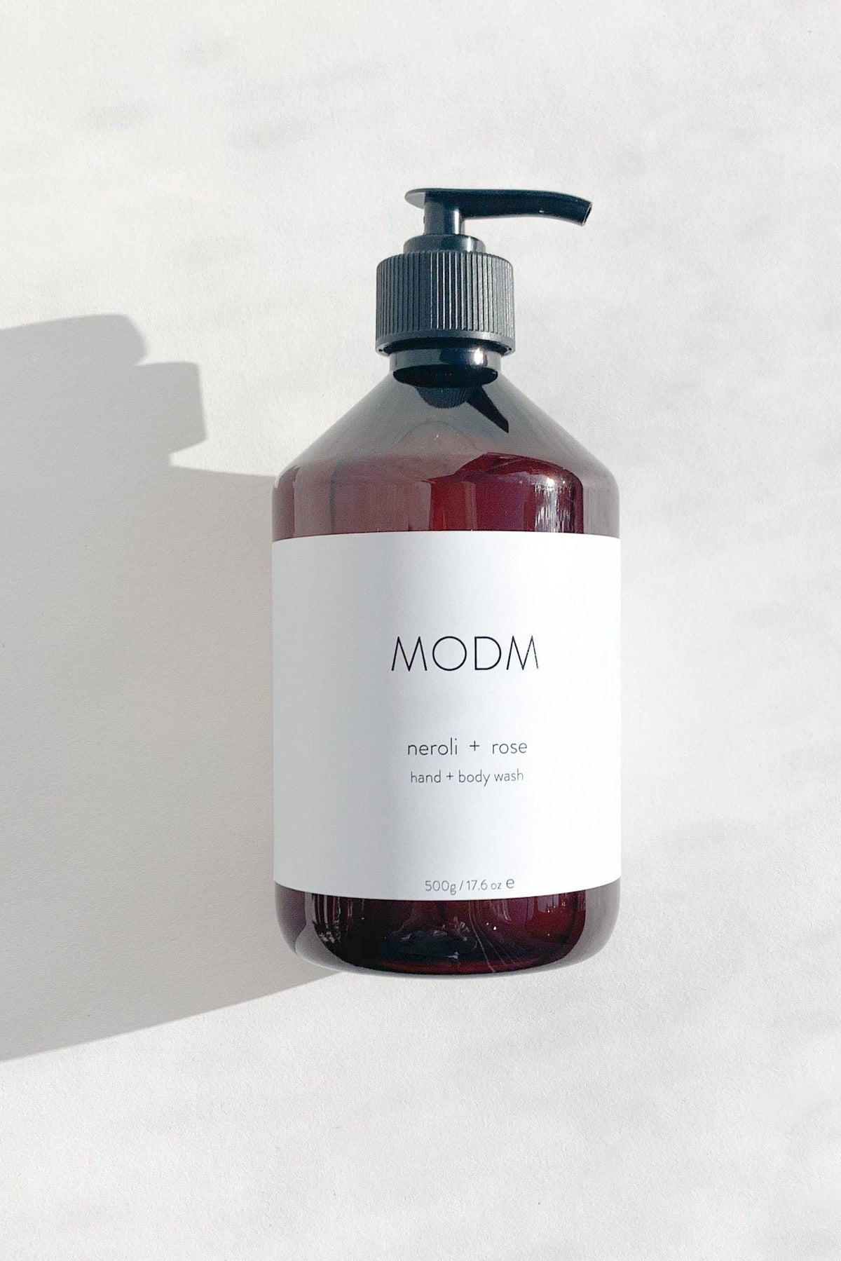 A dark amber bottle of MODM Neroli + Rose hand and body wash with a black pump dispenser and a white label, set against a light gray textured background with a soft shadow.