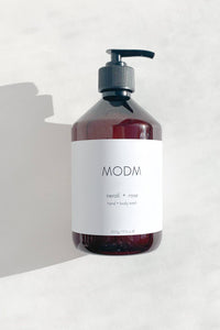A dark amber bottle of MODM Neroli + Rose hand and body wash with a black pump dispenser and a white label, set against a light gray textured background with a soft shadow.
