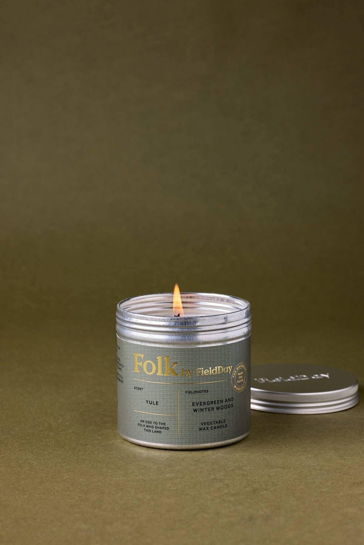 A lit silver metal candle tin with a white label reading 'Folk by FieldDay' sits on a textured olive green surface, with its lid resting beside it.