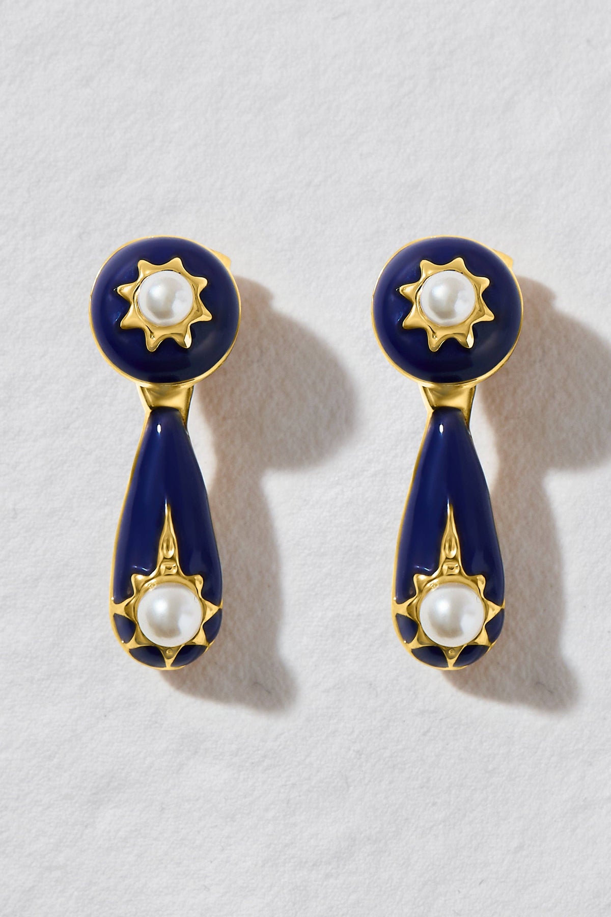 A pair of gold and navy blue drop earrings with white pearl accents, displayed on a textured white background.