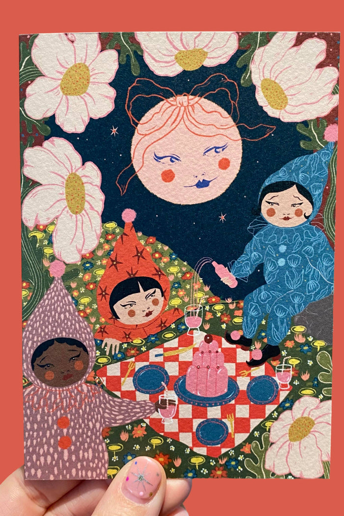 A hand holds a square art print or card featuring a whimsical illustrated scene with three stylized children around a picnic blanket under a dark night sky, surrounded by large white flowers. The print is centered against a solid coral-red background.