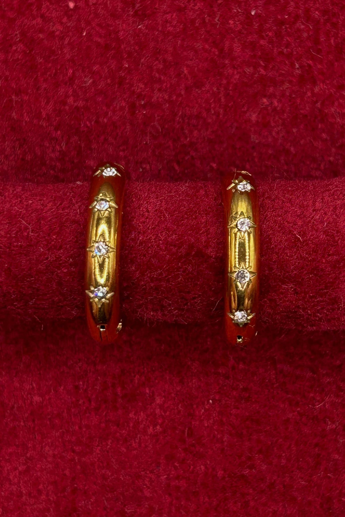 A pair of gold hoop earrings with small clear stones, displayed on a red velvet ring stand.