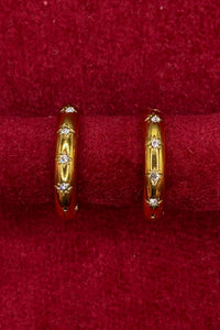 A pair of gold hoop earrings with small clear stones, displayed on a red velvet ring stand.