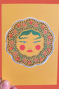 A hand holds a textured mustard yellow art print or card featuring a stylized face framed by a floral mandala border, set against a solid coral-red background.