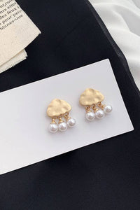 Gold cloud-shaped earrings with pearl drops on a white card against a black fabric background.
