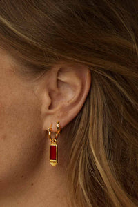 A close-up of a model's ear wearing a gold huggie hoop earring with a small rectangular red gemstone pendant.