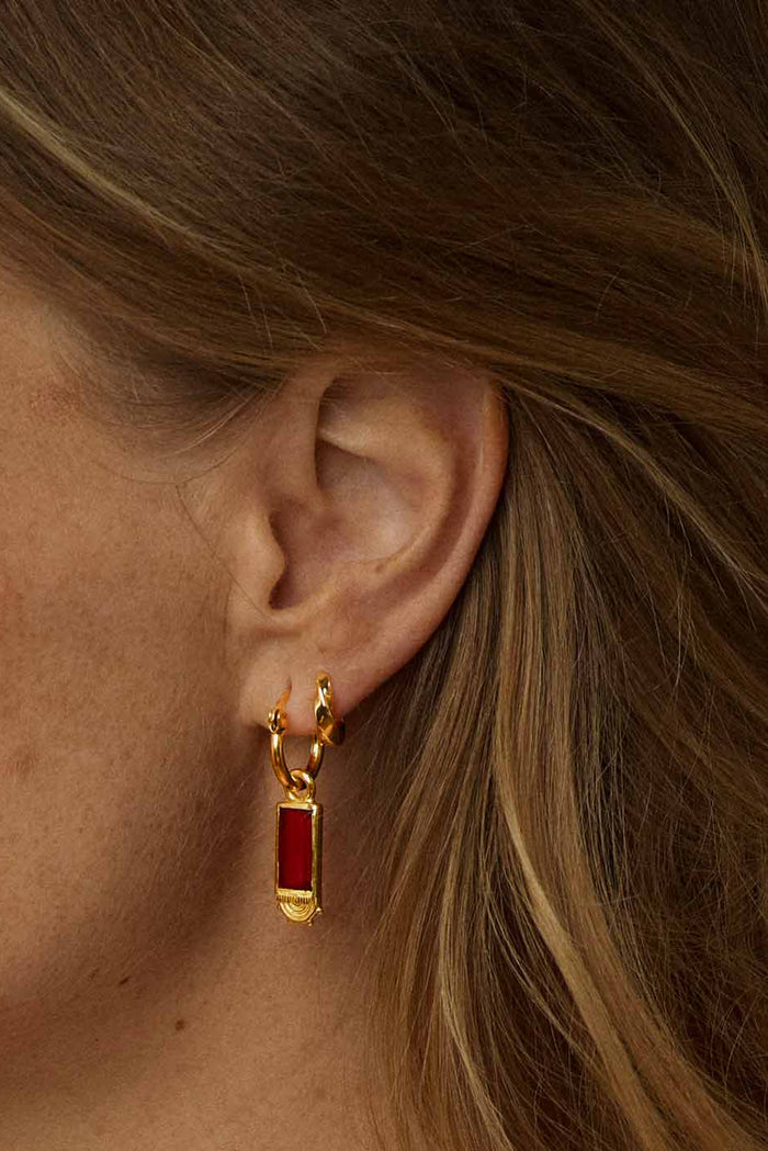 A close-up of a model's ear wearing a gold huggie hoop earring with a small rectangular red gemstone pendant.
