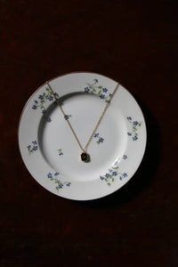 A delicate gold chain necklace with a small dark square pendant, draped over a white ceramic plate decorated with blue flowers, set against a dark brown wooden background.