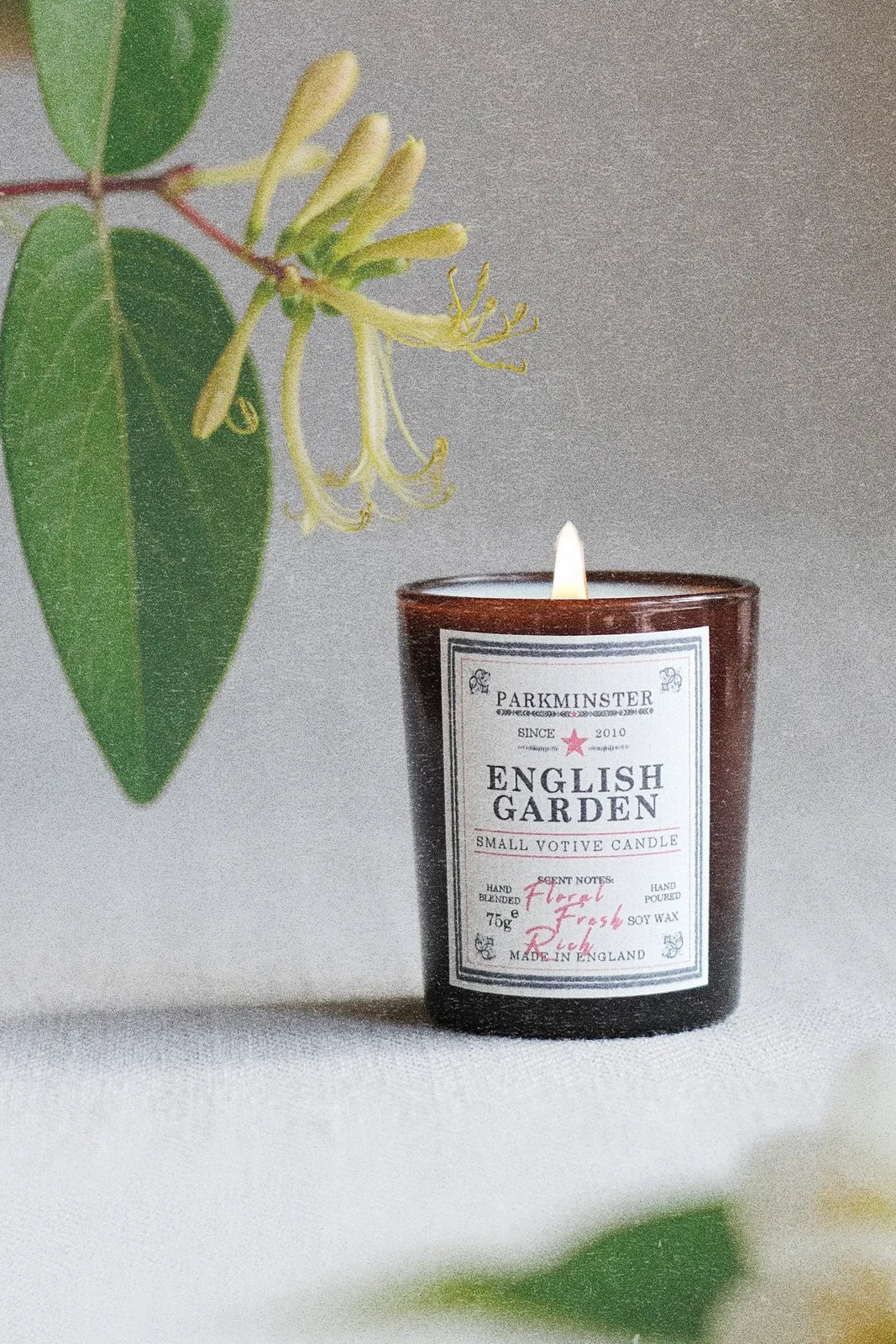 A small amber glass candle jar labeled 'PARKMINSTER ENGLISH GARDEN' with a white wick, styled on a light surface with green leaves and pale flowers in the background.