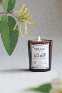 A small amber glass candle jar labeled 'PARKMINSTER ENGLISH GARDEN' with a white wick, styled on a light surface with green leaves and pale flowers in the background.