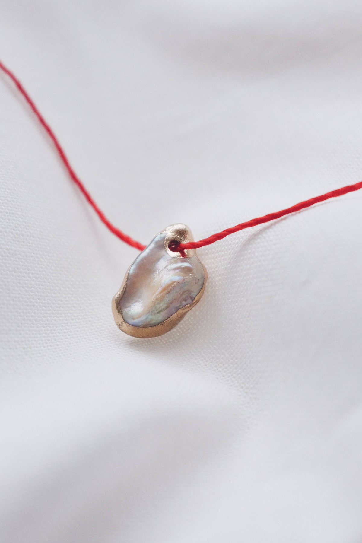 A delicate red cord necklace featuring a small, organic-shaped pendant made from a polished iridescent shell, resting on a soft white fabric background.
