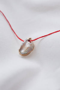 A delicate red cord necklace featuring a small, organic-shaped pendant made from a polished iridescent shell, resting on a soft white fabric background.