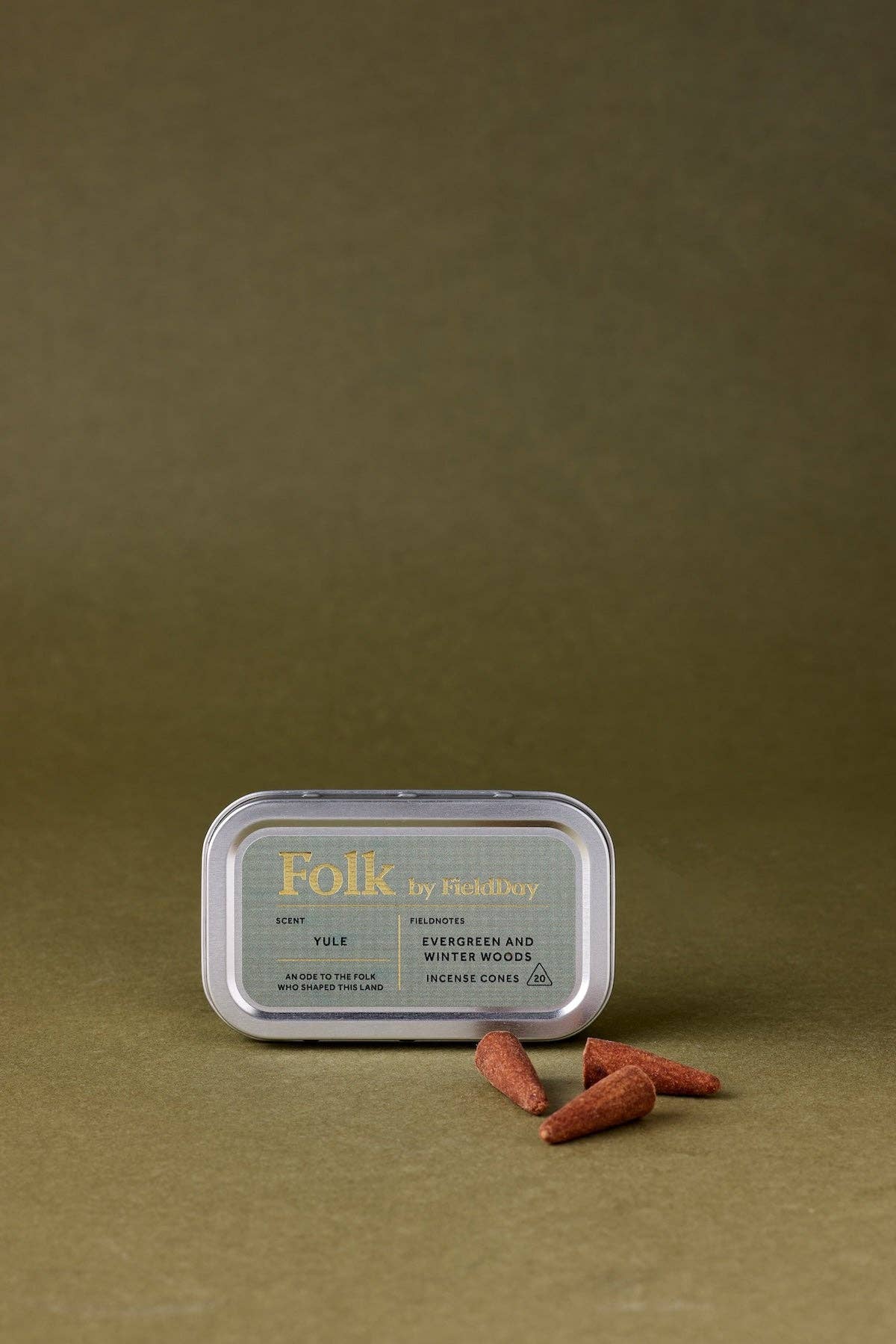 A silver metal tin of Folk by Fjæløy tea with a green label, placed on a textured olive-brown surface with three loose tea bags in front.