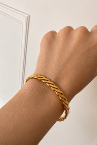 A close-up of a person's wrist wearing a gold braided cable bracelet against a white paneled wall background.
