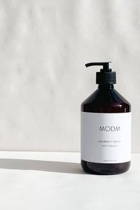 A dark amber glass pump bottle of MODM mandarin + velvety hand & body wash with a white label, set on a white surface against a textured white wall.