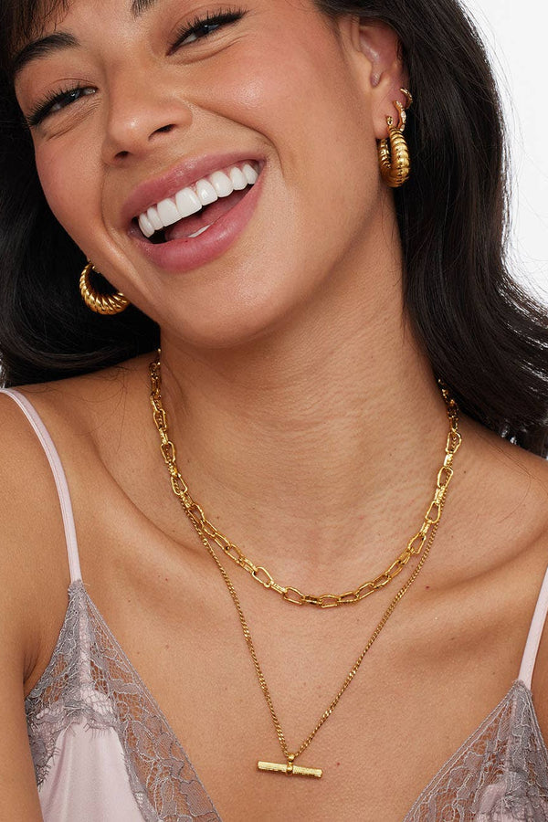 A close-up lifestyle shot of a smiling woman wearing layered gold necklaces, including a curb chain, a cable chain, and a delicate chain with a toggle clasp pendant, along with gold hoop earrings.