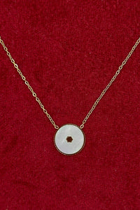 A delicate gold chain necklace featuring a circular white stone pendant with a small central cutout, displayed on a textured red velvet background.