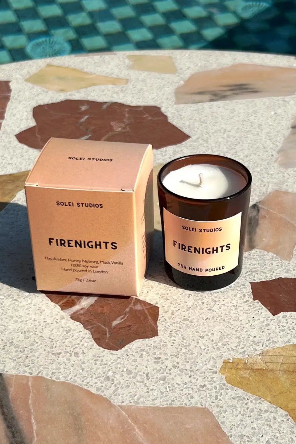 A Solet Studios 'Fire Nights' soy candle in an amber glass jar sits next to its peach-colored cardboard packaging box on a terrazzo-patterned stone table near a swimming pool.