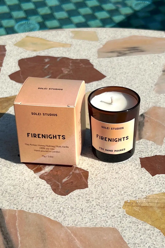 A Solet Studios 'Fire Nights' soy candle in an amber glass jar sits next to its peach-colored cardboard packaging box on a terrazzo-patterned stone table near a swimming pool.