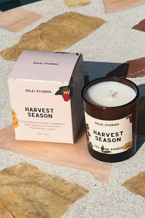 A Soleri Studios 'Harvest Season' candle in a dark amber glass jar next to its light pink packaging box, placed on a stone-tiled surface outdoors.