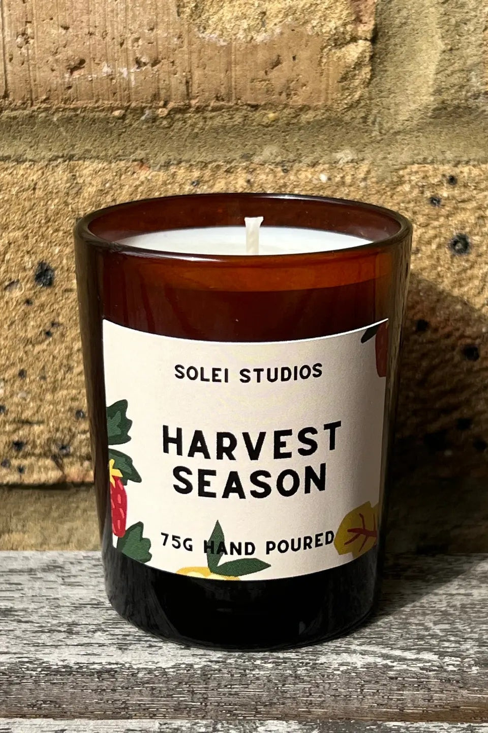 A 75g hand-poured soy candle in an amber glass jar with a white wick, featuring a white label that reads 'SOLEI STUDIOS HARVEST SEASON' with botanical illustrations. The candle is placed on a weathered wooden surface against a rustic tan brick wall.