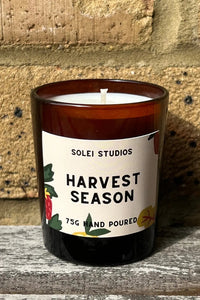 A 75g hand-poured soy candle in an amber glass jar with a white wick, featuring a white label that reads 'SOLEI STUDIOS HARVEST SEASON' with botanical illustrations. The candle is placed on a weathered wooden surface against a rustic tan brick wall.