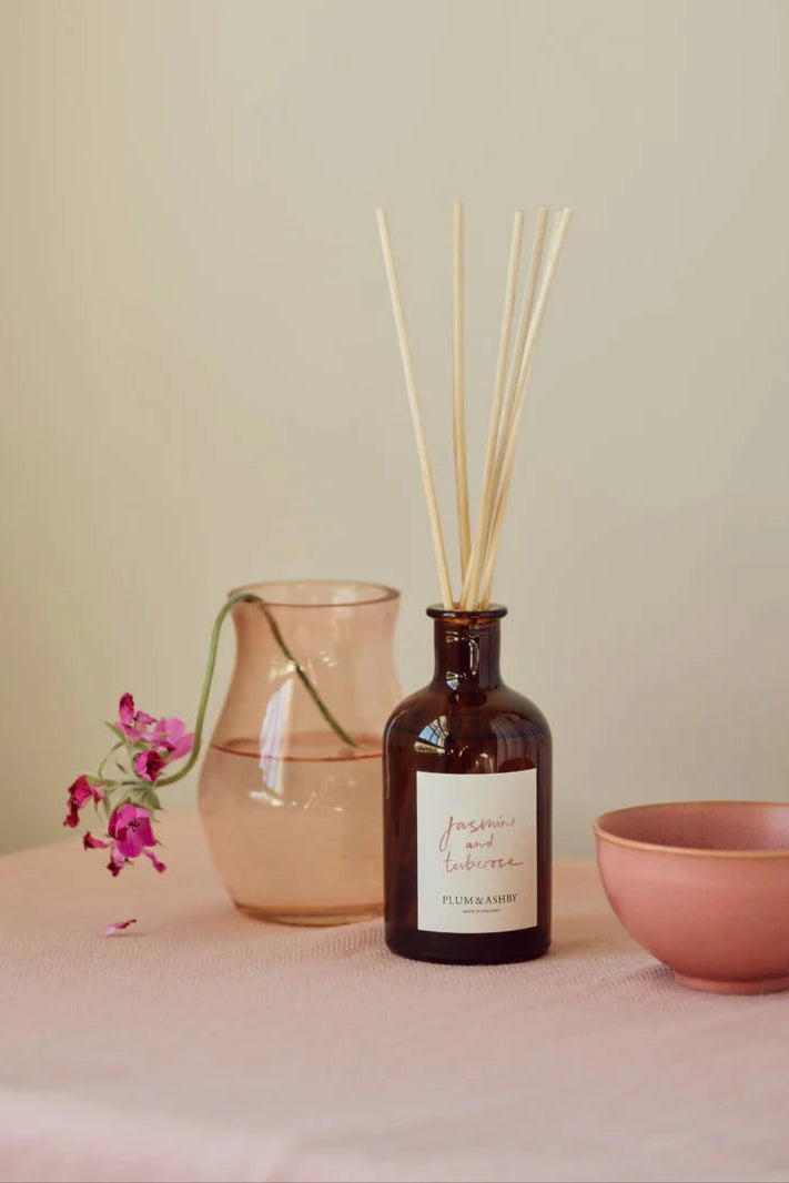 A bottle of diffuser with reeds, a pink vase, and a bowl on a light surface.