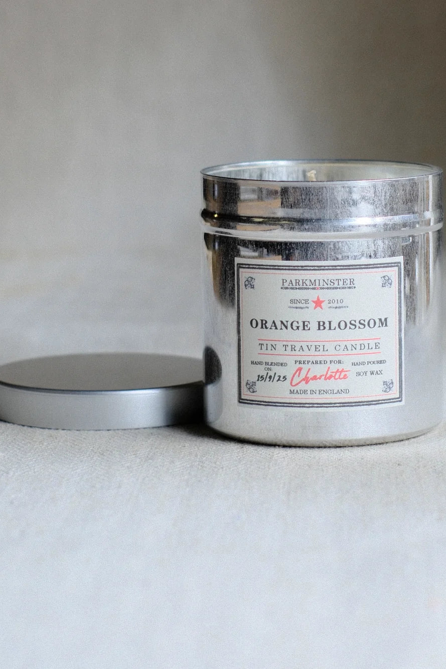 A silver tin travel candle labeled 'Orange Blossom' by Darkminster, with its lid resting beside it on a neutral light gray surface.