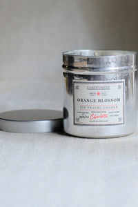 A silver tin travel candle labeled 'Orange Blossom' by Darkminster, with its lid resting beside it on a neutral light gray surface.