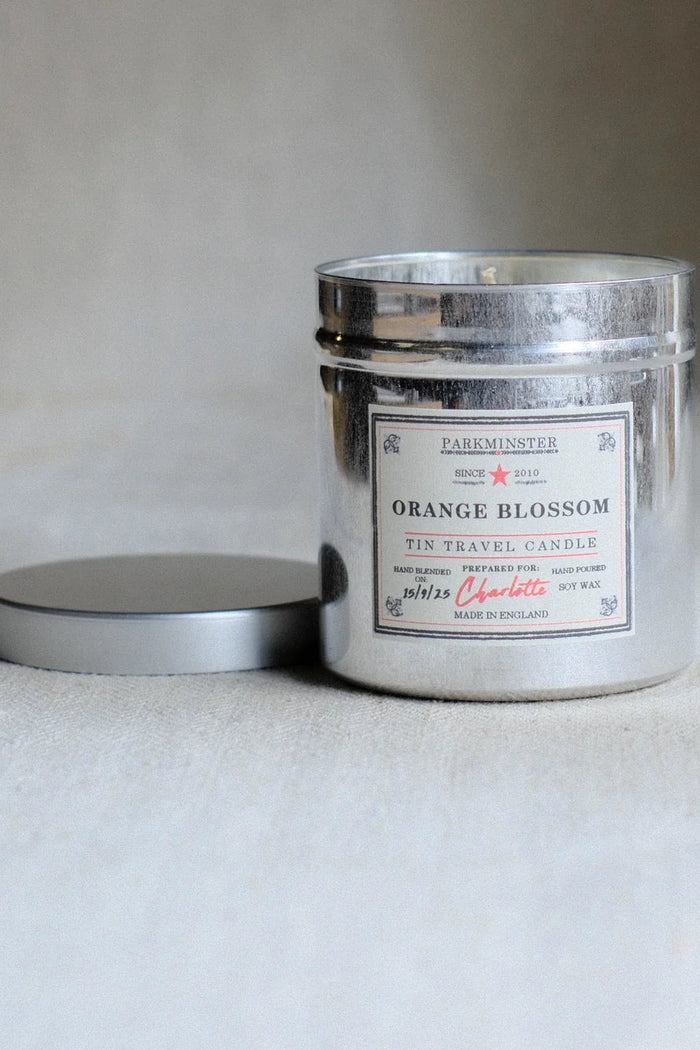 A silver tin travel candle labeled 'Orange Blossom' by Darkminster, with its lid resting beside it on a neutral light gray surface.