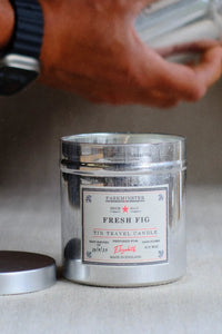 A silver metal tin candle labeled 'Fresh Fig' by Farmminster sits on a light surface with its lid beside it. A person's hand holding a glass container is blurred in the background.