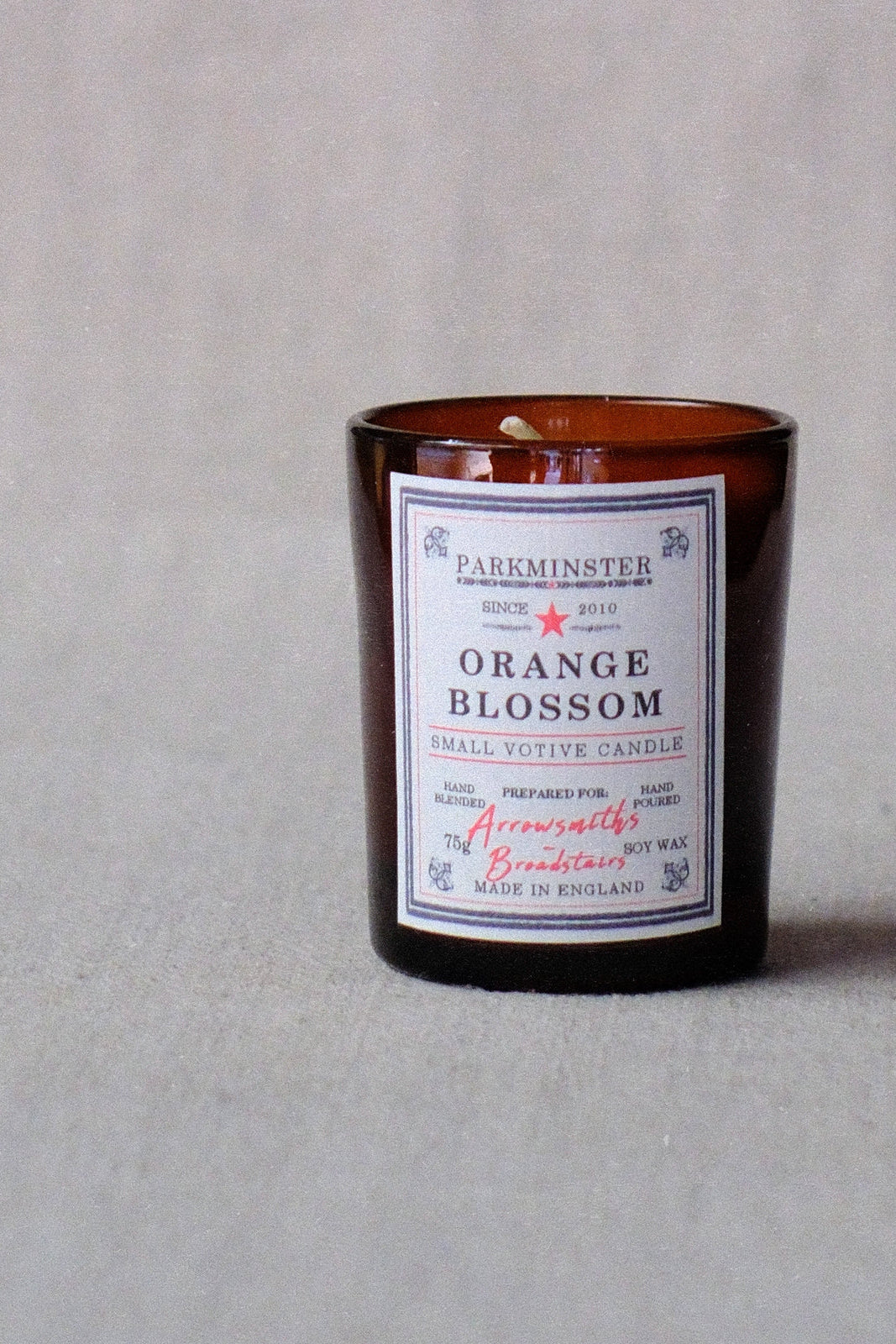 An amber glass jar candle with a white label reading 'PARKMINSTER ORANGE BLOSSOM' and 'SMELL VOTIVE CANDLE', photographed on a light gray textured background.