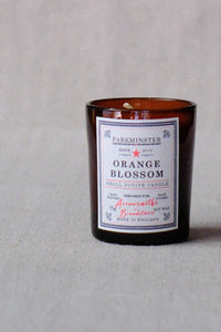 An amber glass jar candle with a white label reading 'PARKMINSTER ORANGE BLOSSOM' and 'SMELL VOTIVE CANDLE', photographed on a light gray textured background.