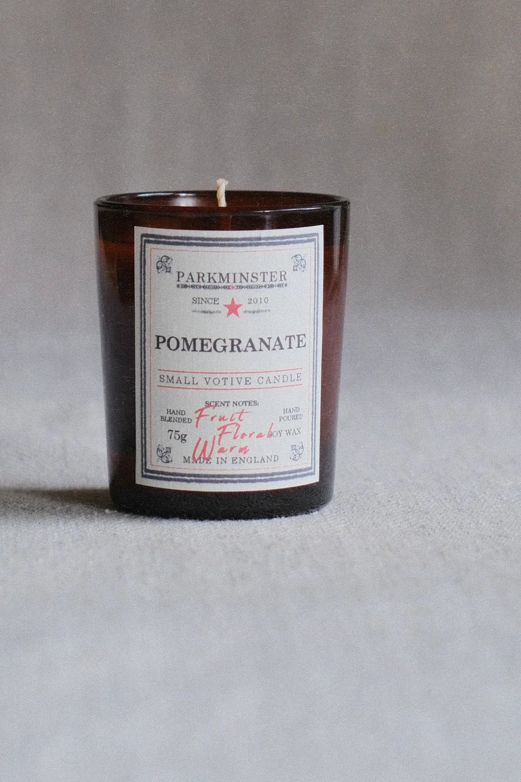 A small amber glass candle jar with a white label reading PARKMINSTER and POMEGRANATE, photographed against a soft gray background.