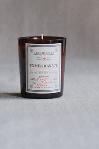 A small amber glass candle jar with a white label reading PARKMINSTER and POMEGRANATE, photographed against a soft gray background.