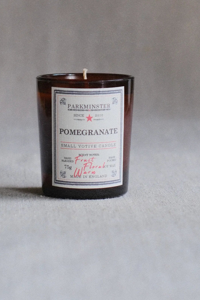 A small amber glass candle jar with a white label reading PARKMINSTER and POMEGRANATE, photographed against a soft gray background.