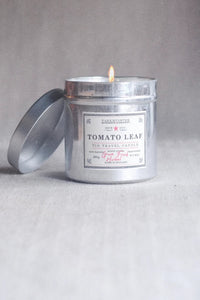 A lit silver metal travel candle tin with its lid off, labeled 'Tomato Leaf' by Parthenon, set against a neutral gray background.