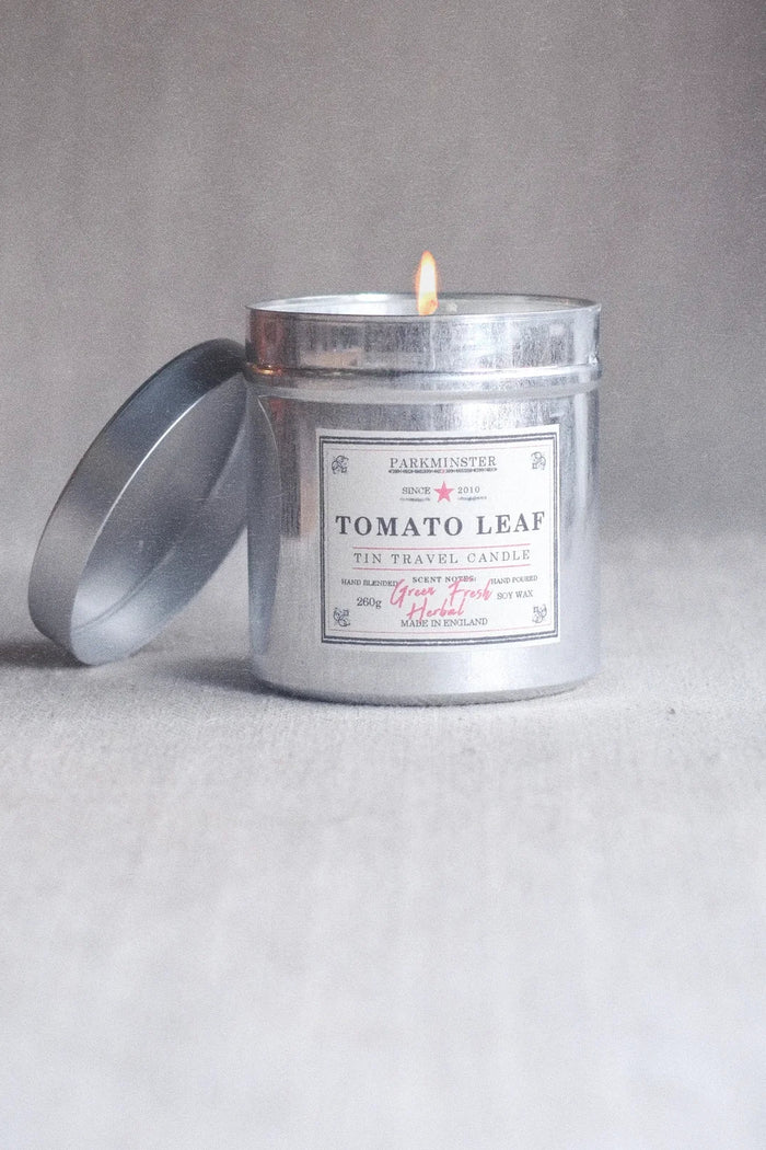 A lit silver metal travel candle tin with its lid off, labeled 'Tomato Leaf' by Parthenon, set against a neutral gray background.