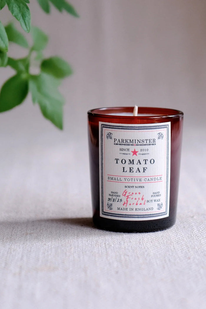 A small amber glass candle jar labeled PARKMINSTER TOMATO LEAF, placed on a light fabric surface with blurred green leaves in the background.