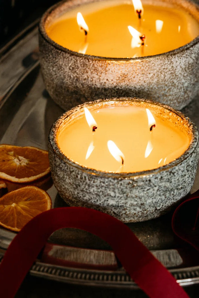 Two lit candles in textured gray ceramic holders sit on a silver tray with dried orange slices and a red ribbon, in a warm, cozy setting.
